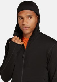 Zwarte zip-up jas met een aansluitende capuchon, voorzien van duimgaten in de mouwen en een contrasterende oranje binnenkant. Glad materiaal met subtiele naden.