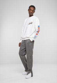 Hvit langermet genser med trykte sleeve-flagg, kombinert med grå avslappede bukser og hvite lavtopp joggesko.