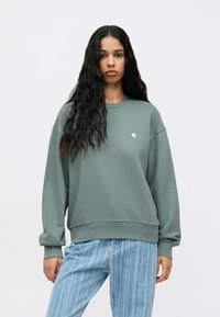 Zöld kapucnis pulóver hosszú ujjakkal, kerek nyakkivágással és bordázott mandzsettákkal. Kicsi logó található a mellen. Világos csíkozott farmerral párosítva.