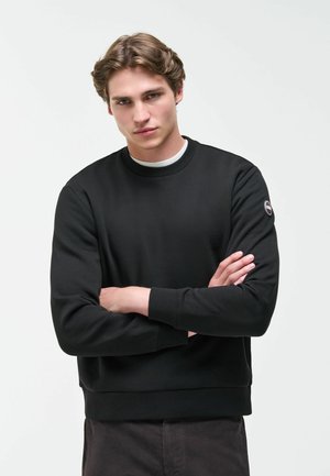 Junger Mann mit hellbraunen Haaren, der einen schwarzen Sweatshirt und dunkle Hose trägt, steht mit verschränkten Armen vor einem weißen Hintergrund.