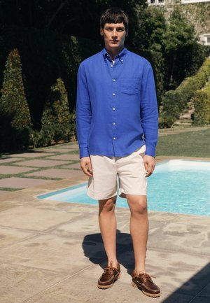 Mann steht neben dem Pool und trägt ein blaues Hemd mit Knöpfen, weiße Shorts und braune Bootsschuhe in einem sonnigen Gartensetting.