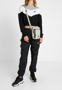 Svart, vit och grå croppad sweatshirt med logotyp; svarta texturerade joggers med sidoknappar; ljusgrå axelväska med gula accentfärger.