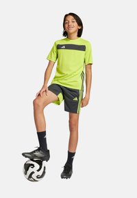 Camiseta atlética de color amarillo neón con detalles en negro, pantalones cortos grises con ribetes neones, zapatillas de fútbol negras y un balón de fútbol en blanco y negro.