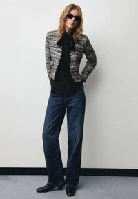 Zwart-wit gestructureerde tweedjas, zwarte blouse, wijde blauwe spijkerbroek en zwarte haklaarzen, gedragen met oversized donkergekleurde zonnebril.