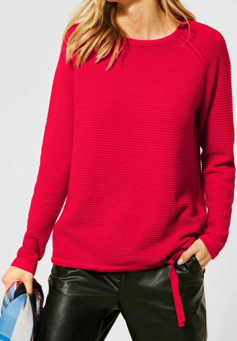 Femme portant un pull rouge vif texturé à manches longues avec cordon de serrage à l'ourlet et un pantalon en cuir noir, tenant un tissu à carreaux bleu dans une main.