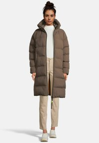 Brauner Puffercoat mit Kapuze, Reißverschluss vorne, gesteppter Textur und seitlichen Taschen. Kombiniert mit einem cremefarbenen Pullover und hellen Hosen.