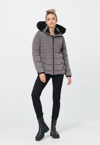 Graue Pufferjacke mit einer schwarzen Fellkapuze, ausgestattet mit einem Reißverschluss und horizontalem Steppmuster. Schwarze Jeans und klobige schwarze Stiefel vervollständigen den Look.