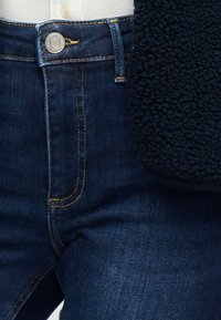 Κοντινό πλάνο στη μέση ενός σκούρου μπλε τζιν με κουμπί και κίτρινα ραφτά, συνδυασμένο με υφασμένο μανίκι μαύρου σακακιού.