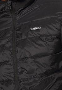 Czarna kurtka puchowa z pikowanym wzorem, wyposażona w przedni zamek błyskawiczny i oznaczoną metkę. Gładki, błyszczący materiał o lekkiej fakturze.