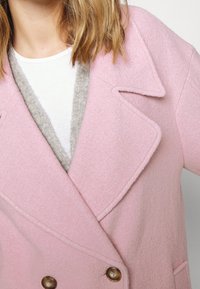 Doppelreihiger rosa Wollmantel mit hellbraunen Knöpfen, übergroßen Revers und einem strukturierten grauen Innenkragen, der unter einem weißen Hemd hervorlugt.