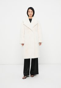 Faux fur frakke i blød hvid, med bred revers, lange ærmer og lige snit. Kombineret med sorte wide-leg bukser og strop sandaletter.