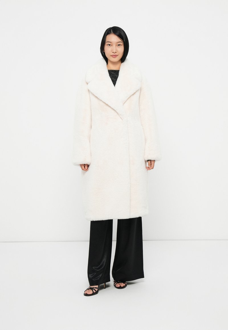 Faux fur frakke i blød hvid, med bred revers, lange ærmer og lige snit. Kombineret med sorte wide-leg bukser og strop sandaletter.