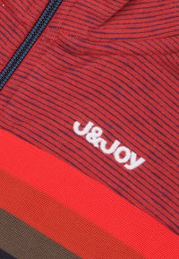 Rood gestreepte stof met marineblauwe accenten en een wit "J&JOY" logo. Bevat een ritssluiting en textuurpatronen in horizontale lijnen.