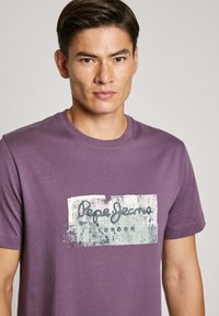 Violetinės spalvos medvilnės marškinėliai su grafiniu spaudiniu, kuriame užrašyta "Pepe Jeans London" ant tekstūruoto fono. Trumpomis rankovėmis ir apvalia iškirpte.