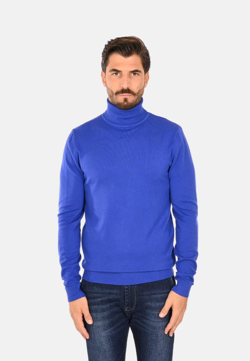 Maglione a collo alto blu, texture a costine, maniche lunghe e design aderente. Indossato con jeans in denim scuro su uno sfondo neutro.
