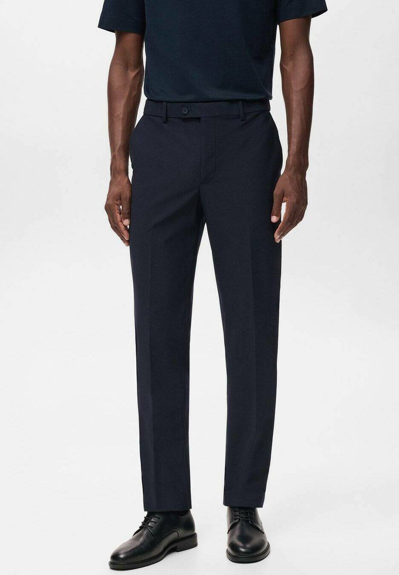 Pantalons de costume bleu marine avec une coupe slim, devant plat et deux poches latérales. Le tissu a une texture lisse, associé à des chaussures noires à lacets.
