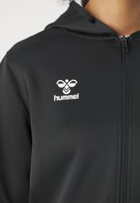 Must zip-up kapuutsiga dressipluus valge hummel logoga vasakul rinnal, kantud isiku poolt, kellel on nähtav kael ja lokkis juukseotsad.