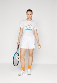 Hvit tennisantrekk som består av en t-skjorte med logo og shorts, kombinert med oransje sokker, hvite joggesko, et tennisracket og en gul ball.