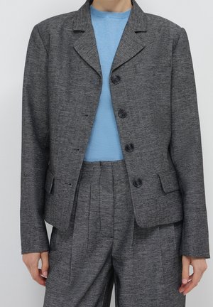 Person iført en skræddersyet grå blazer med fiskebenmønster og højtaljede matchende bukser over en lyseblå sweater med rund hals.