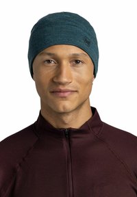 Young man wearing a teal beanie and a burgundy zip-up sweater, facing forward with a neutral expression on a white background.