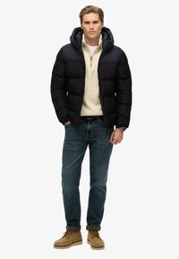 Czarna kurtka puchowa z kapturem, z zamkiem z przodu, noszona na jasnobeżowym swetrze. Połączona z niebieskimi dżinsami i beżowymi butami. Minimalistyczny design.