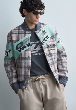 Homme portant une veste varsity à carreaux avec des panneaux turquoise et un texte graphique noir, un T-shirt graphique foncé, un pantalon beige à cordon de serrage et des lunettes de soleil noires.