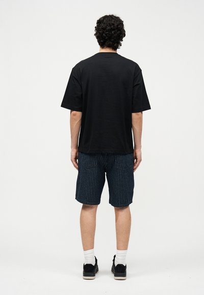 Άνδρας με σγουρά μαλλιά στέκεται με την πλάτη γυρισμένη, φορώντας μια μαύρη φαρδιά μπλούζα, ναυτικό σορτς με ρίγες, άσπρες κάλτσες και μαύρα αθλητικά παπούτσια.