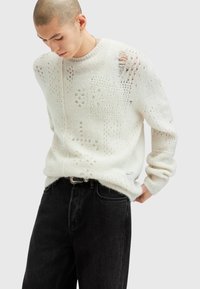 Weißer, gestrickter Pullover mit einem offenen Strickmuster und überschnittenen Schultern, kombiniert mit schwarzen, eng anliegenden Jeans und einem Gürtel.