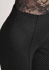 Close-up van een persoon die zwarte broek met kleine witte stippen en een zwart kanten top draagt.