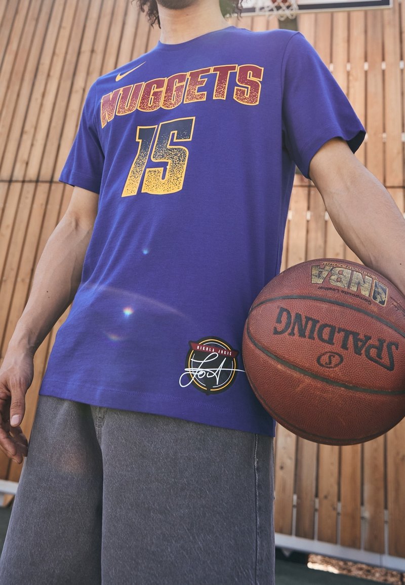 Personne en maillot violet des Denver Nuggets et short gris tenant un ballon de basket Spalding NBA en plein air près d'une clôture en bois.