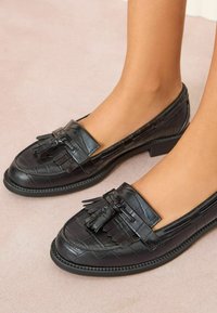 Mustmustest nahast loafers, millel on tekstureeritud krokodillimuster, dekoratiivne frillide aksent ja madal, kihiline kannaosa.