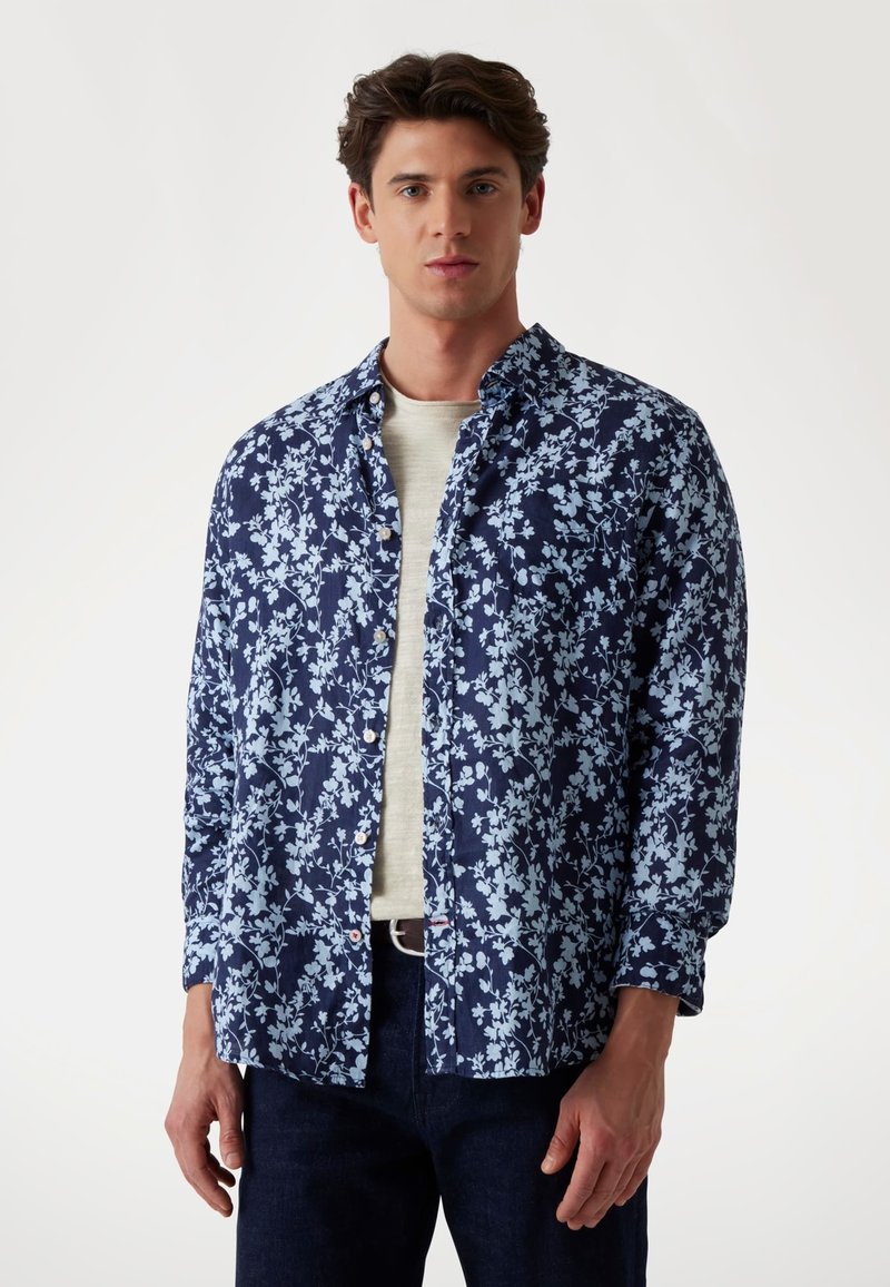 Camicia blu navy con un leggero motivo floreale, design con bottoni, maniche lunghe e vestibilità rilassata, indossata sopra una t-shirt grigia chiara.