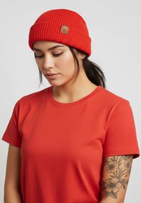 Red ribbed beanie with a leather tag, worn with a matching red t-shirt. The fabric appears soft, with a snug shape and texture.