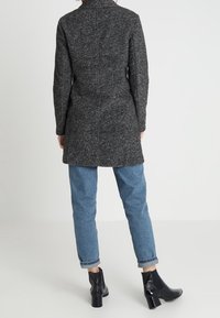 Cappotto in misto lana grigio scuro con colletto strutturato, dettagli della cucitura posteriore e maniche lunghe. Indossato con jeans azzurri con risvolto e stivaletti neri.