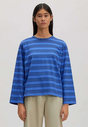 Vrouw staat met het gezicht naar voren, draagt een blauw shirt met lange mouwen met zwarte horizontale strepen en beige broek tegen een effen achtergrond.