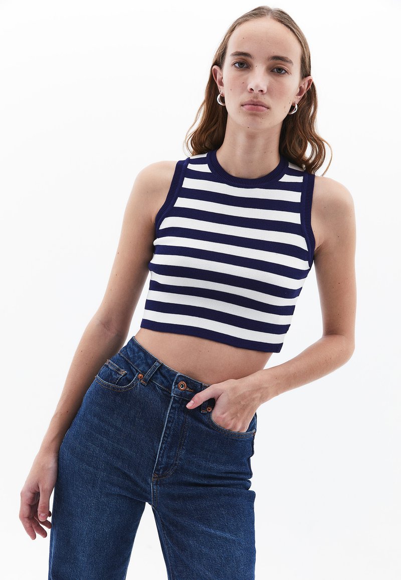 Top corto a righe in blu navy e bianco, texture a costine, design senza maniche, abbinato a jeans a vita alta di un blu scuro. Stile casual e aderente.