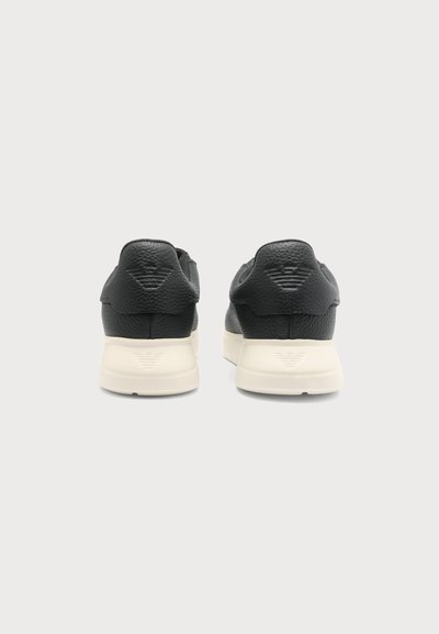 Paire de baskets en cuir noir avec semelles blanches vues de dos sur fond blanc, avec un logo embossé subtil.