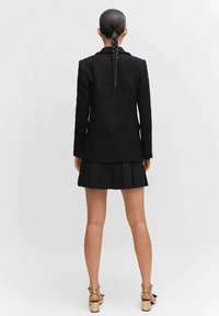 Blazer negro de tweed con un corte entallado, combinado con una falda negra con pliegues. La modelo posa con un peinado trenzado y sandalias de tacón color bronceado.