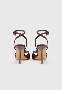 Brune stiletto hæle med krydsede stropper, åben tå design, glat tekstur og en tynd, forlænget hæl. Har en guldfarvet spænde detalje.