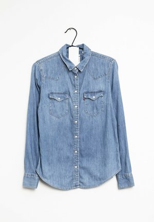 Jasnoniebieska dżinsowa koszula z długim rękawem, zapinana na guziki, z dwoma kieszeniami na piersi, wisząca na czarnym wieszaku na białym tle.