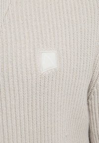 Gai bēša adīta džemperis ar ribotu tekstūru, kuram kreisajā pusē ir neliels taisnstūrveida logotipa uzlīme ar smalku dizainu.