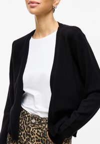 Μαύρο κασκόλ φτιαγμένο από απαλό υλικό, με ανοιχτό μπροστινό σχέδιο και ριμπ μανίκια, συνδυασμένο με λευκή μπλούζα και παντελόνι με μοτίβο λεοπάρ.