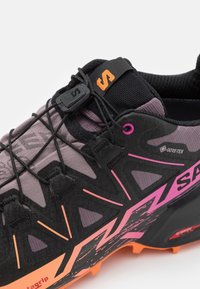 Gros plan d'une chaussure de course noire et violette avec des accents roses, système de laçage rapide, étiquette Gore-Tex et semelle orange.