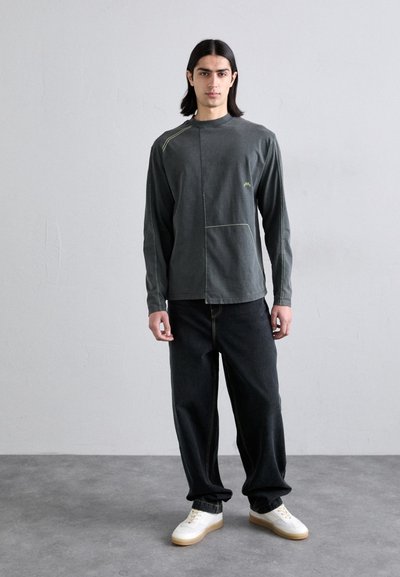 Chemise à manches longues, verte foncée avec des coutures jaunes contrastantes et une coupe large, associée à un jean noir ample et des baskets blanches.