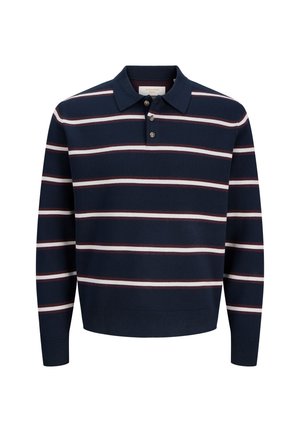 Jeune personne debout portant un polo à manches longues rayé bleu marine, un pantalon marron et des chaussures beiges, regardant légèrement sur le côté et souriant.