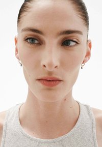 Pendiente de aro dorado con un acento circular, mostrado en un primer plano del rostro de una persona; piel clara, ojos azules, vistiendo una blusa gris con textura en relieve.