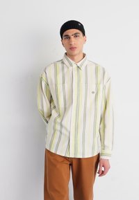 Camisa de botón a rayas en verde claro, amarillo y blanco; corte relajado; dos bolsillos en el pecho; combinada con pantalones marrones y un gorro de punto negro.