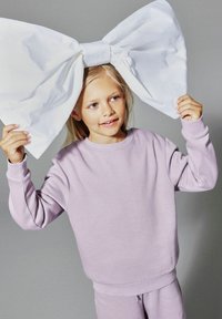 Fille blonde portant un sweat-shirt lilas et un pantalon tient un grand nœud blanc au-dessus de sa tête sur un fond gris.