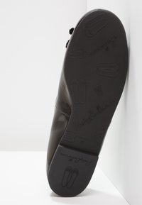Semelle de chaussure noire avec ballerines embossées et logo "PrettyBallerinas" visible, positionnée verticalement contre une surface blanche.