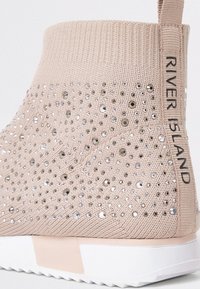 Beige ankelstövel i ett texturerat stickat material, dekorerad med silverfärgade rhinestones, med en märkt etikett och en vit sula.